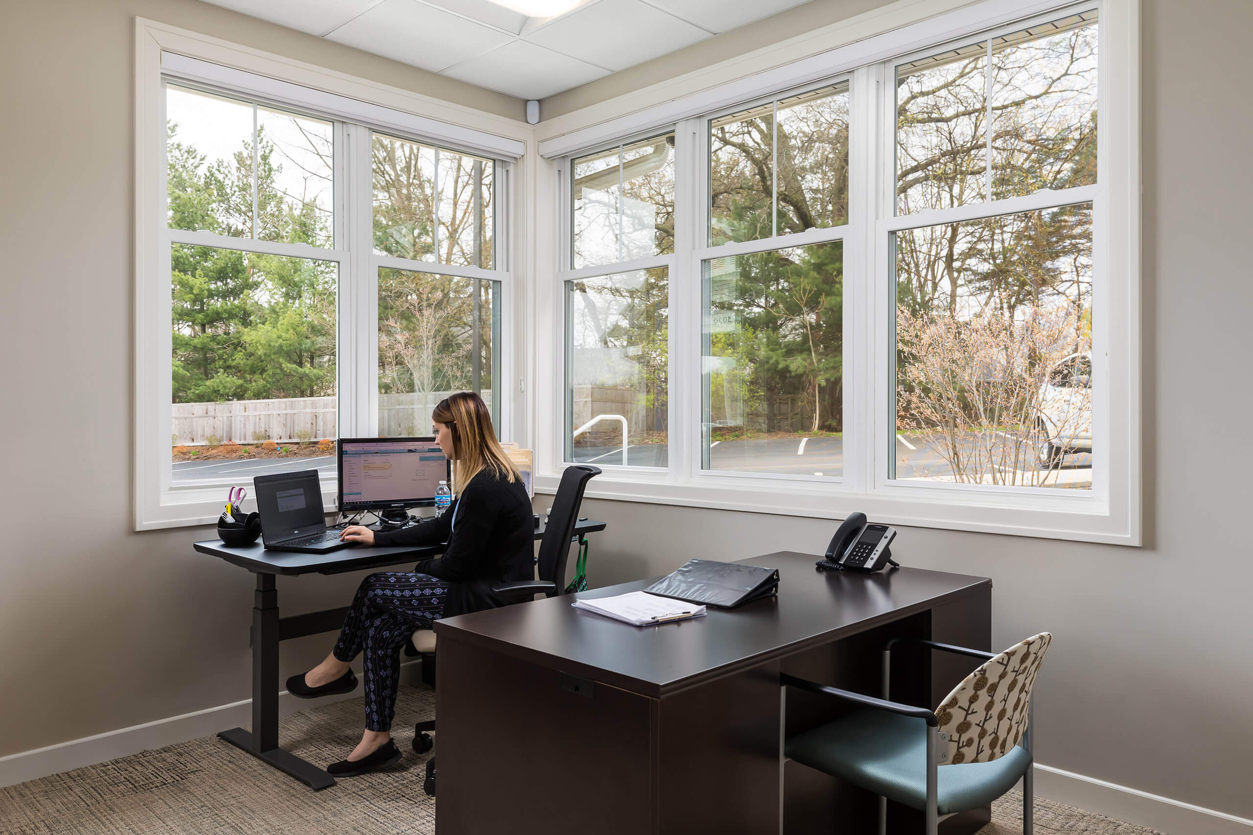 A woman sitting at a black standing desk in the corner of an office with windows