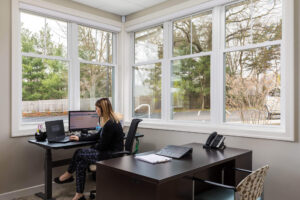 A woman sitting at a black standing desk in the corner of an office with windows