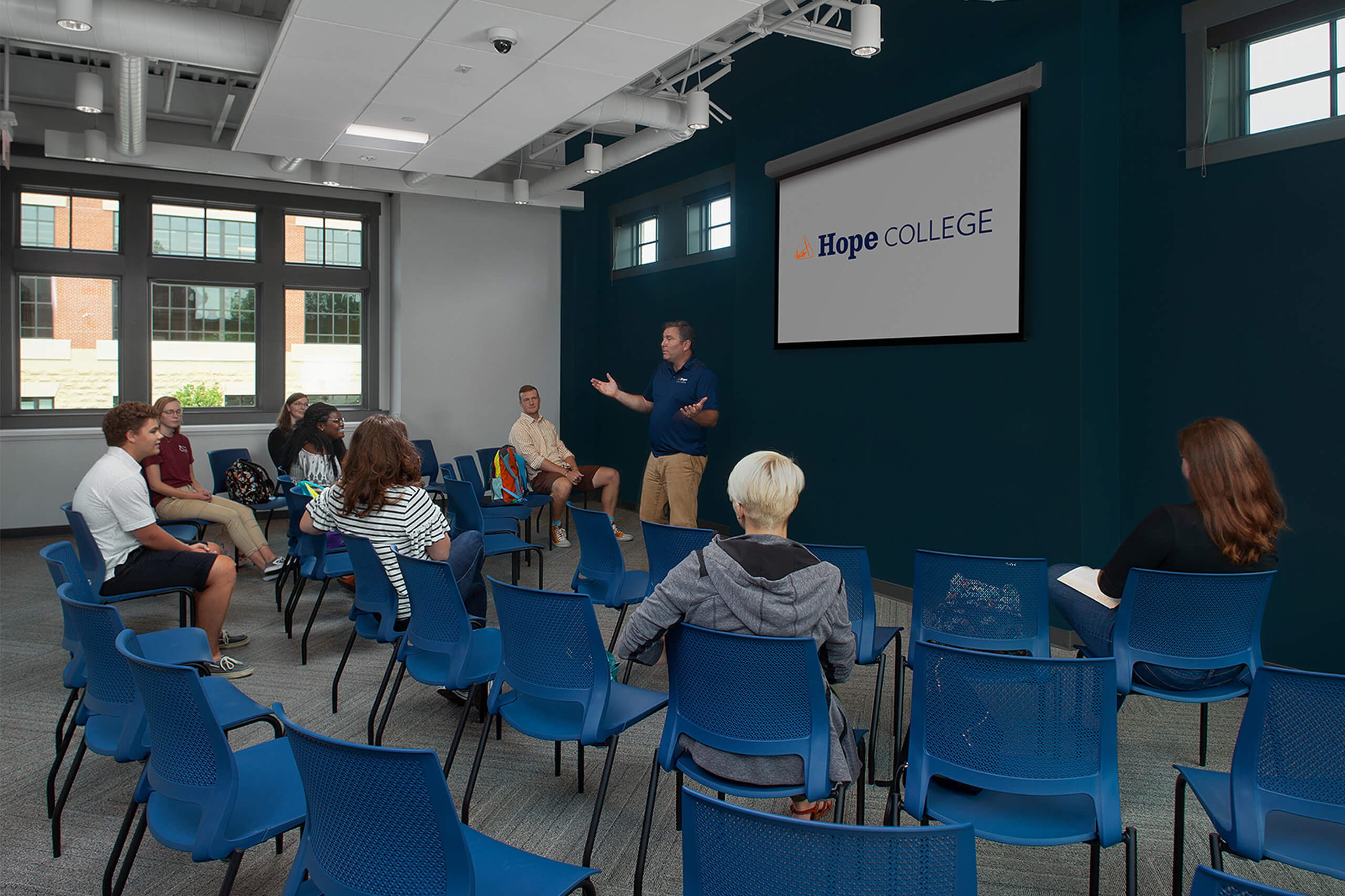 Professor stand in front of a Hope College banner speaking to eight students seated in rows of blue chairs.