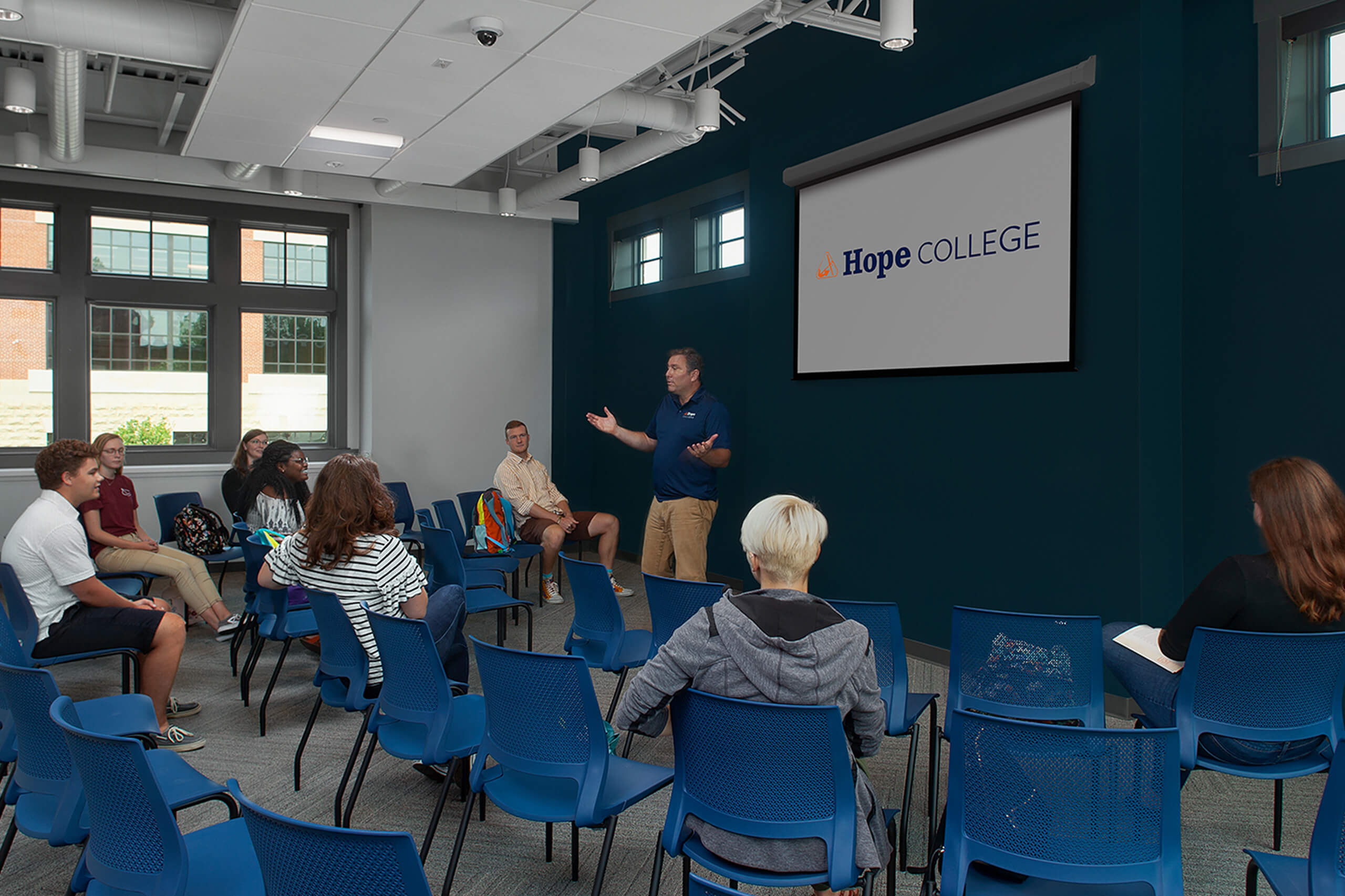 Professor stand in front of a Hope College banner speaking to eight students seated in rows of blue chairs.