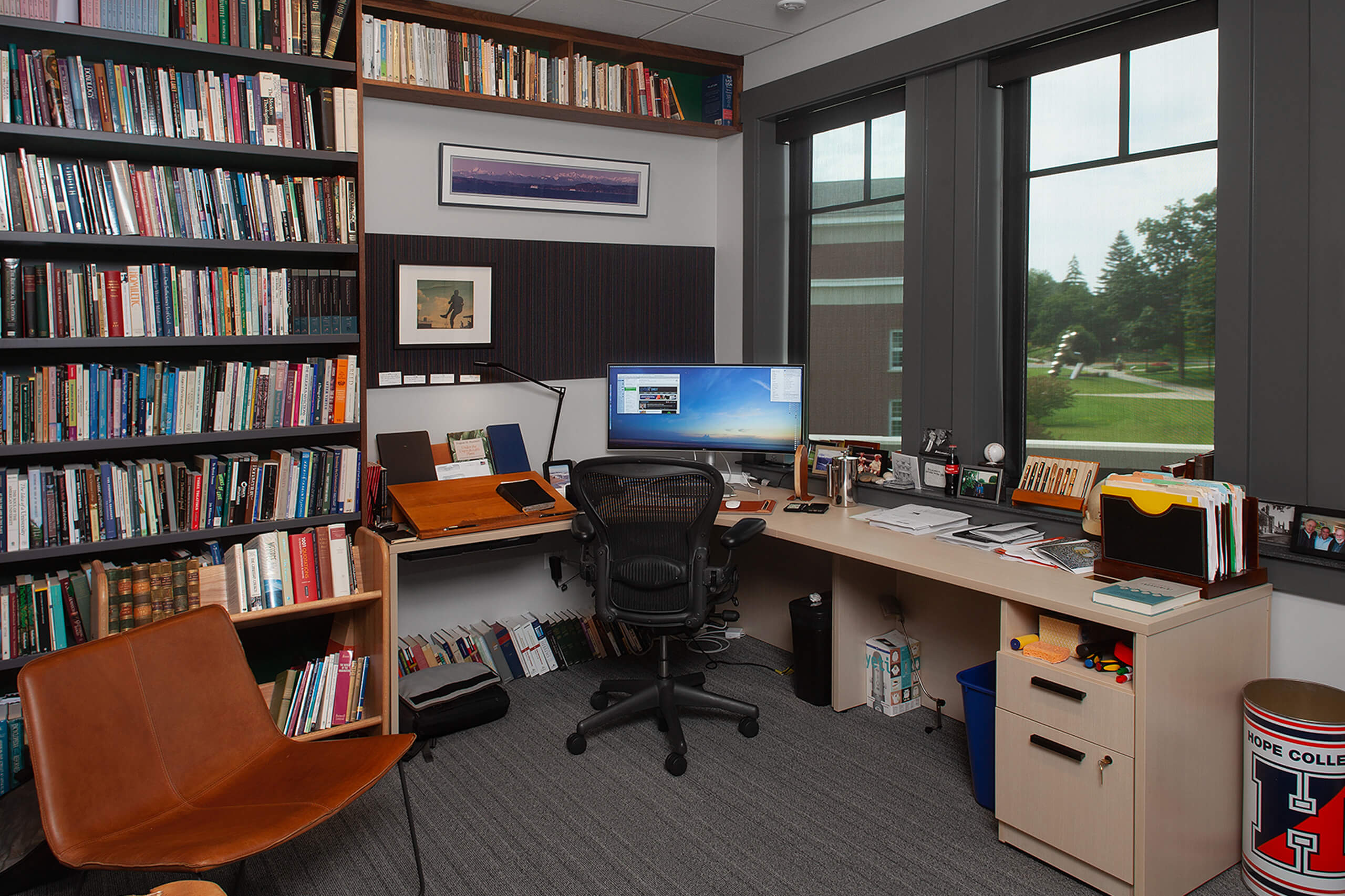 Professors office with a full bookcase and l-shaped desk covered in notebooks, files and a desktop computer.