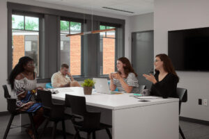 Four students in a meeting room, three of them are seated at a table with a tv behind them the other is seated in a chair along the wall.