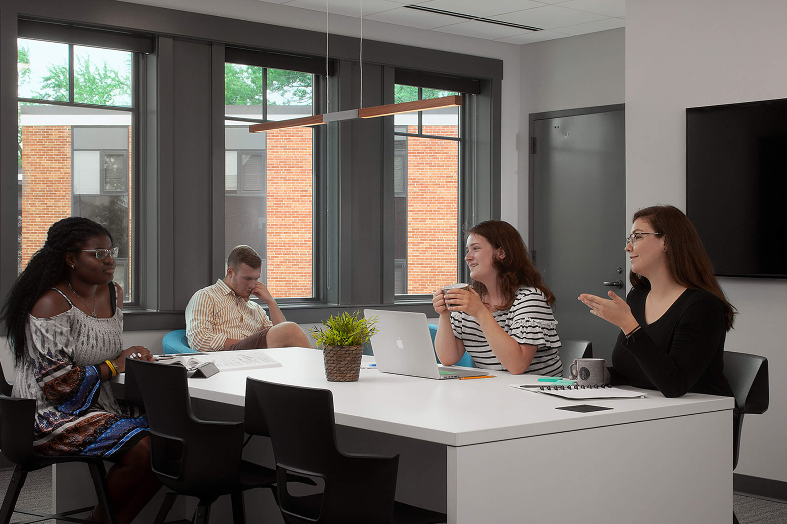 Four students in a meeting room, three of them are seated at a table with a tv behind them the other is seated in a chair along the wall.