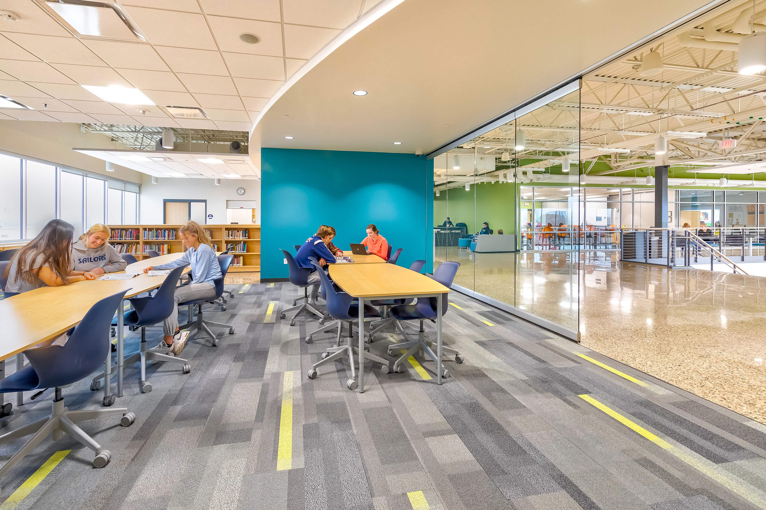 Students sitting at rectangular tables learning in a bright room with a teal wall and a glass wall