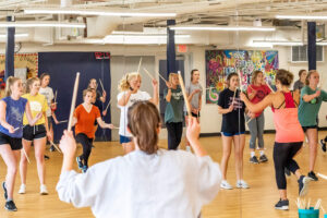 Students standing and holding drumsticks in a room with a wall mirror