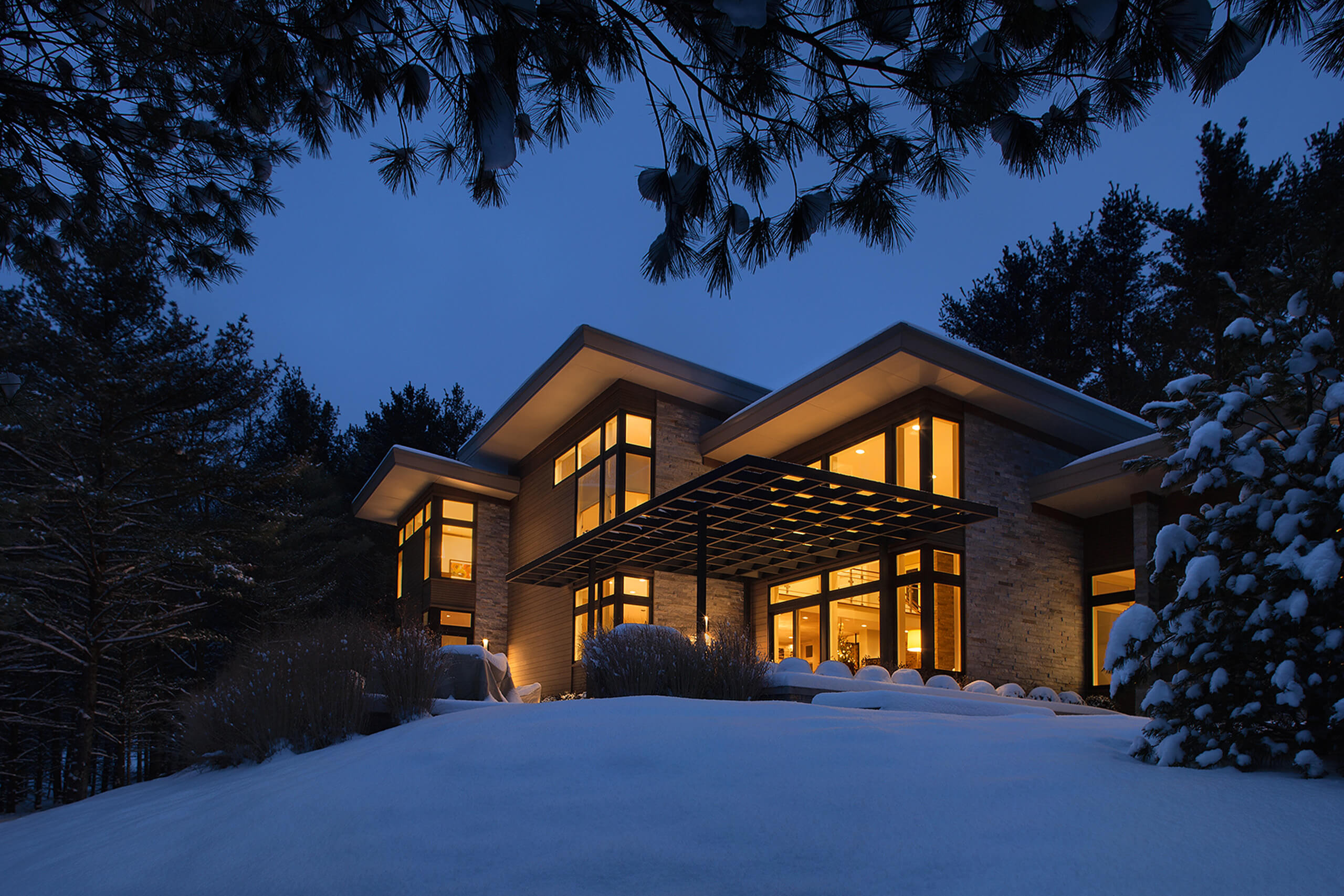 exterior view of a modern residential home at night lit from within with snow covering the ground and pine trees