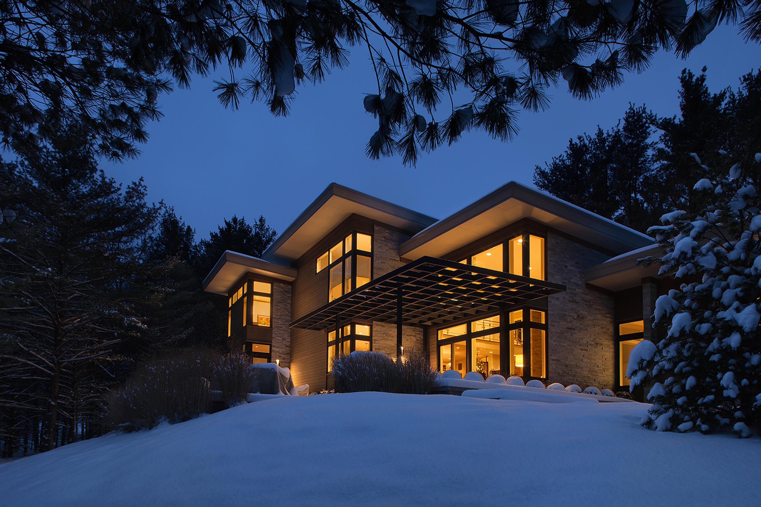 exterior view of a modern residential home at night lit from within with snow covering the ground and pine trees