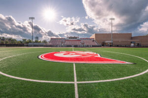 The logo in the middle of the football field with the bleachers in the background at Trojan Stadium