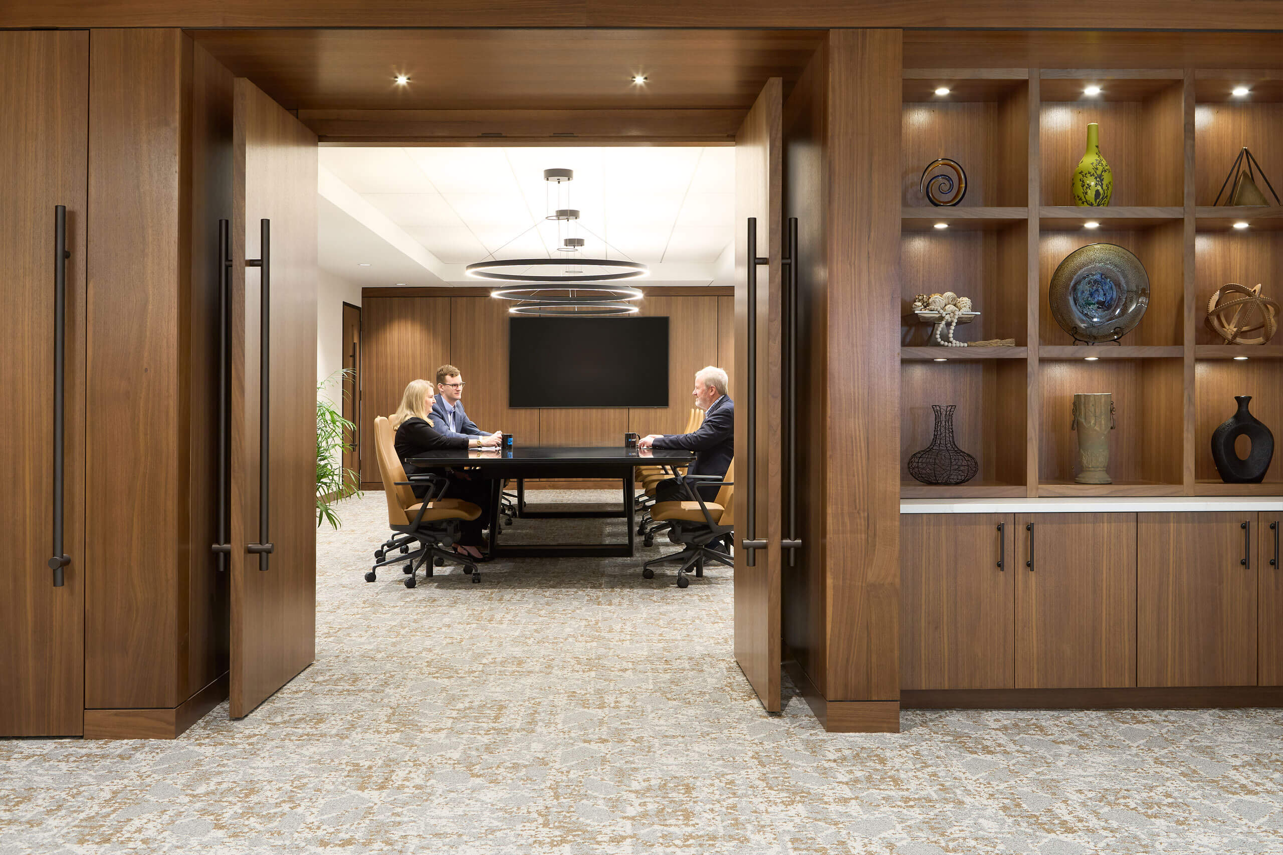 interior view of a modern office workspace with dark wood paneled doors facing a boardroom with people sitting at the table