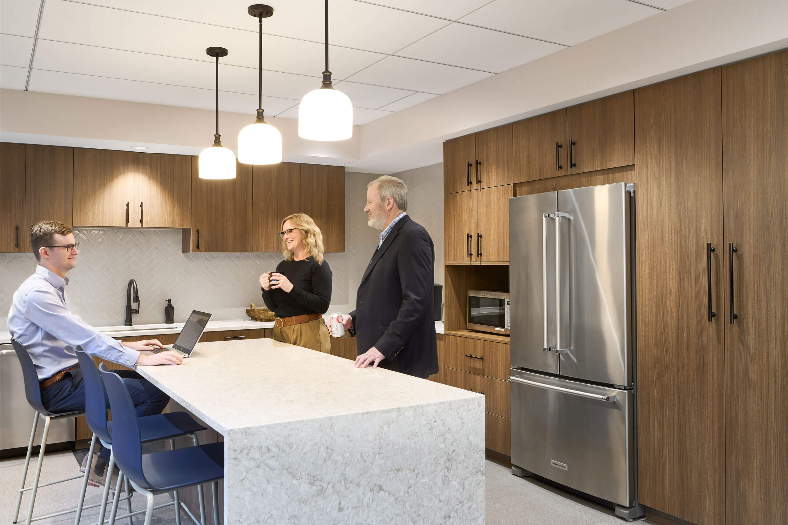 3 coworkers interact in an interior kitchen of an office space with a kitchen island and barstools