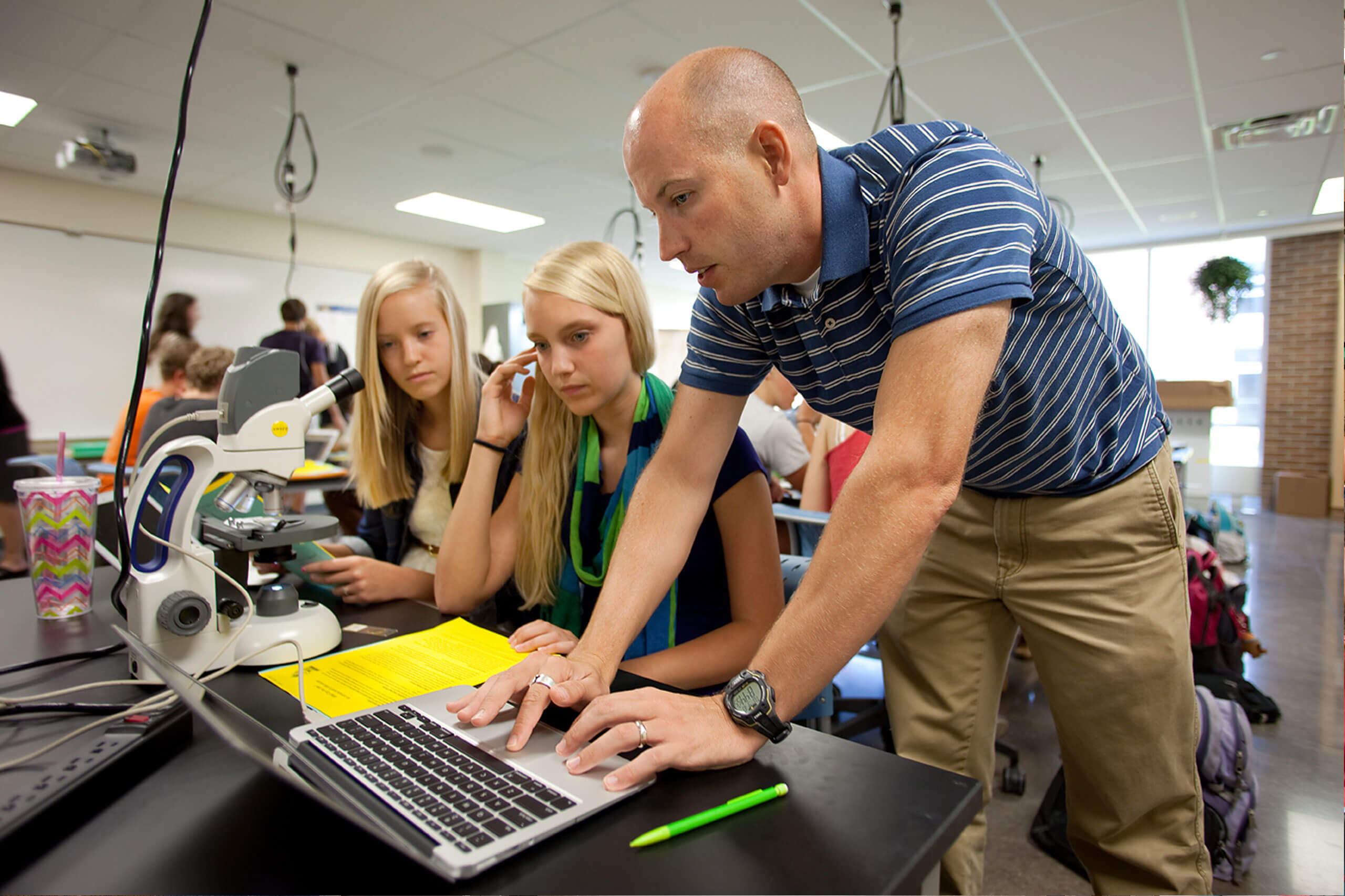 Grand Rapids Christian High School male science teacher showing two female students his laptop screen while they use a microscope