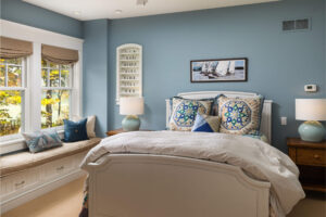 interior bedroom of a shingle-style cottage with a double bed with colorful pillows and a window seat with cushions